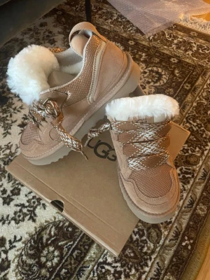 Bruna sneakers från UGG med fuskpäls - Säljer ett par beige sneakers från UGG med grova snören i brun och vitt. Skorna har en fluffig vit fuskpäls på insidan och detaljer i mocka och mesh. Sulan är tjock och beige, perfekt för kalla dagar. UGG-logga på sidan och bak. Säljer pga de var för små. De är i 40 men känns som 39