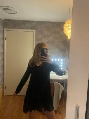 Svart långärmad klänning från Amisu - Säljer en svart klänning från Amisu med v-ringning och långa ärmar. Klänningen har en luftig och volangig nederdel som ger en skön och avslappnad känsla. Perfekt för dig som gillar enkel och stilren design med lite extra flow.