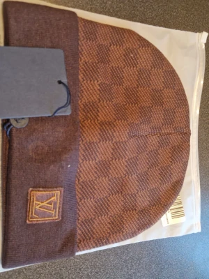 Brun rutig mössa från Louis Vuitton - Snygg brun mössa från Louis Vuitton med rutigt mönster och LV-logga framtill. Mössan har en bred uppvikt kant och är tillverkad i mjukt material som känns skönt mot huvudet. Perfekt för dig som vill ha en stilren accessoar med lyxig känsla.