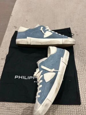 Philippe Model blå sneakers, strl 42 - Säljer ett par Philippe Model sneakers i blått och vitt. Skorna har en snygg låg profil, rund tå och platt sula. Ovandelen är i mocka och skinn med vita snören och detaljer. Kommer med originalpåse och låda. Perfekt för dig som gillar stilrena sneakers.