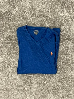 Blå v-ringad t-shirt från Polo Ralph Lauren - Snygg blå t-shirt från Polo Ralph Lauren med v-ringning och klassisk orange logga broderad på bröstet. Tillverkad i mjuk bomull och har korta ärmar. Perfekt till jeans eller shorts för en clean och enkel look. Tröjan är använd så slitage finns 