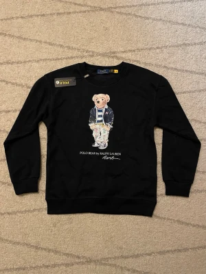 Svart sweatshirt Polo Bear Ralph Lauren - Svart sweatshirt från Polo Ralph Lauren i storlek S med ikoniskt Polo Bear-tryck på bröstet. Rund hals och långa ärmar med ribbade muddar. Materialet är mjuk bomull, perfekt för chill dagar. Snygg och klassisk design med coolt motiv framtill.