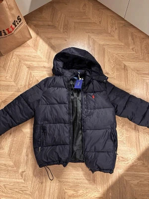 Mörkblå pufferjacka från Polo Ralph Lauren - Säljer en mörkblå pufferjacka från Polo Ralph Lauren med huva och dragkedja. Jackan har två fickor med dragkedja och den klassiska röda loggan på bröstet. Perfekt för kalla dagar och riktigt snygg till jeans eller mjukis.  Säljes för att den var för stor på mig.    Helt ny!                                     