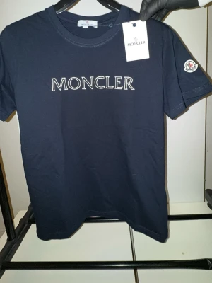 Mörkblå Moncler t-shirt med logga - Snygg mörkblå t-shirt från Moncler i bomull med stor vit Moncler-logga på bröstet och ett broderat Moncler-märke på ärmen. Klassisk rund hals och korta ärmar, perfekt för dig som gillar stilrena designer och exklusiva märken.