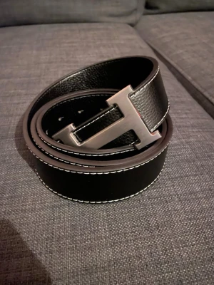 Svart skärp med H-spänne - Snyggt svart skärp med ett stort H-format metallspänne. Skärpet har vit kontrastsöm längs kanterna och är gjort i ett slätt, strukturerat material som ger en lyxig känsla. Perfekt för att lyfta din outfit med en stilren detalj.