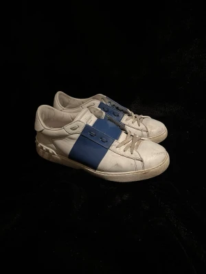 Valentino Open sneakers vit/blå - Säljer ett par Valentino Open sneakers i vitt skinn med bred blå rem över mitten. Skorna har rund tå, snörning och platt sula med ikoniska nitar baktill. Perfekt för dig som gillar stilrena och lyxiga sneakers med en cool twist. Skorn är smått slitna därefter priset ! Cond 7/10 fullt använd bara 