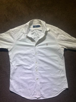 Vit skjorta från Ralph Lauren M - Säljer en klassisk vit skjorta från Ralph Lauren i storlek M. Skjortan har knappar hela vägen, krage och den ikoniska broderade loggan på bröstet. Materialet är mjuk bomull och modellen har normal passform med lång ärm. Defekt längst fram vid ärmen, syns ej om man har nått över!