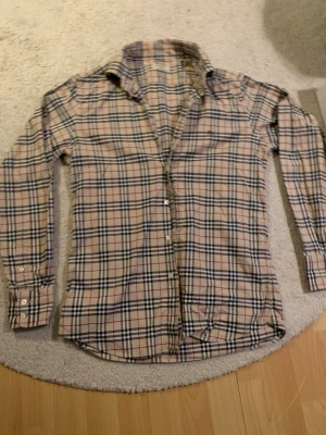 Burberry skjorta - Säljer en klassisk Burberry skjorta i slim fit med det ikoniska rutiga mönstret i beige, svart, vit och röd. Skjortan har sin själv unika still som kan passa till nästan vad som helst. 