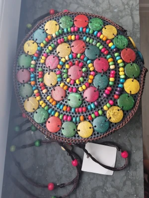 Rund axelväska med färgglada pärlor - Unik rund axelväska med färgglada pärlor och plattor i grönt, gult, rött och blått. Väskan har brunt virkat material runt kanterna och detaljerade snören med pärlor. Stängs med dragkedja och har ett bohemiskt, kreativt uttryck som sticker ut. Aldrig använt feån kreta. Nypris 399kr pris kan diskuteras