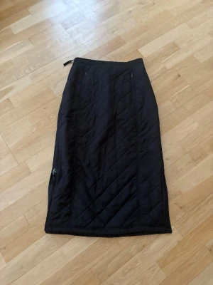 Svart quiltad långkjol från Clique - Quiltad svart kjol från Clique med dragkedjor i sidorna och elastisk midja. Två fickor fram med dragkedja. Kjolen har ett snyggt rutmönster och är tillverkad i syntetmaterial som känns mjukt och värmande. Perfekt för dig som vill ha en stilren och bekväm look under kyliga dagar.