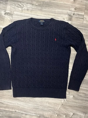 Mörkblå stickad tröja Polo Ralph Lauren - Säljer en mörkblå stickad tröja från Polo Ralph Lauren i storlek barn XL men sitter som storlek herr S. Tröjan har klassiskt kabelstickat mönster, rund hals och en liten röd broderad logga på bröstet. Perfekt för dig som gillar stilrena och tidlösa plagg.