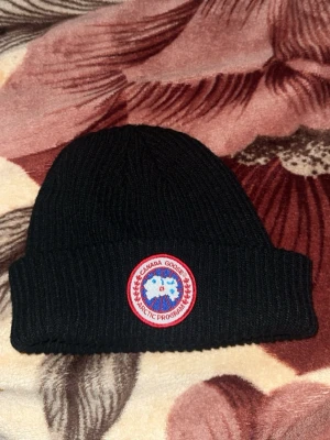  Canada Goose mössa  - Säljer Svart ribbstickad mössa från Canada Goose med klassisk rund logotyp framtill i rött, vitt och blått. Mössan har uppvikt kant och är tillverkad i ett mjukt stickat material som håller värmen. Perfekt för kalla dagar och snygg till streetwear. Mössan är helt ny 