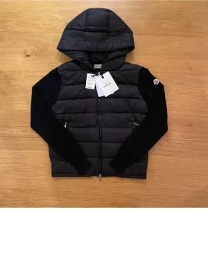 Svart pufferjacka från Moncler med huva - Svart pufferjacka från Moncler med broderad logga på ärmen och huva med tryck. Jackan har långa stickade ärmar, dragkedja framtill och quiltad kropp. Perfekt för dig som vill ha en snygg och varm jacka med exklusiv känsla.