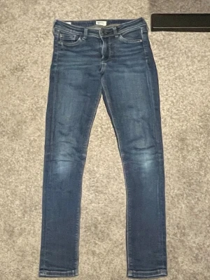 Blå jeans från Pepe Jeans London - Snygga blå jeans från Pepe Jeans London med klassisk femficksdesign och slitna detaljer vid fickan. Jeansen har kontrastsömmar och en brun läderpatch bak i midjan. Modellen har en rak passform och är tillverkad i mjukt denimtyg.