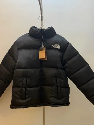 Svart pufferjacka från The North Face - Svart pufferjacka från The North Face i storlek M. Jackan har hög krage, två sidofickor och broderad logga på bröstet och ryggen. Materialet är syntet och dun, med 700-fill power för extra värme. Stängs med dragkedja framtill och har justerbara ärmslut.