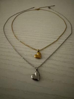 Halsband med hjärthänge i guld och silver( styck pris) - Två stilrena halsband med hjärthängen, ett i guldton och ett i silverton. Kedjorna är tunna och smidiga, och hjärtana har en mjukt rundad form. Perfekt för att bära tillsammans eller var för sig för en enkel men söt look.