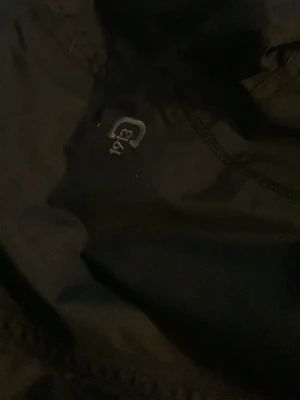 Svart vindjacka från Didriksons1913 - Svart vindjacka från Didriksons1913 med flera fickor, dragkedjor och knappdetaljer. Jackan har en stilren look med broderad logga på bröstet och praktiska detaljer som gör den perfekt för blåsiga dagar. Materialet är slitstark polyester och jackan har långärmade ärmar.