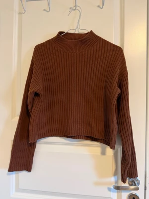Brun ribbad stickad tröja - Säljer en brun stickad tröja med ribbad struktur och hög hals. Tröjan har långa ärmar och en loose passform som ger en chill vibe. Perfekt att matcha med jeans eller kjol för en snygg look.