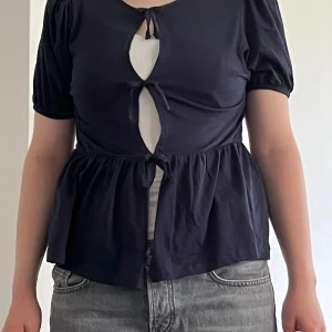 Marinblå blus med knyt från Gina Tricot - Supersöt marinblå blus från Gina Tricot med korta puffärmar och tre knytningar framtill som skapar öppna partier. Modellen har en volangkant nedtill och är tillverkad i mjukt bomullstyg. Perfekt för dig som gillar trendiga detaljer och en feminin look. Vit linne har ej till!