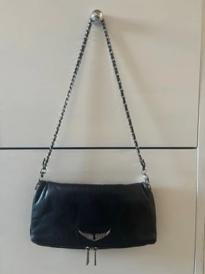 Svart axelväska med kedja från Zadig & Voltaire - Cool svart axelväska i skinn från Zadig & Voltaire med silverfärgad kedja och emblem med vingar framtill. Väskan har dragkedja både fram och bak, vilket gör den praktisk och snygg. Perfekt storlek för det viktigaste man behöver. Säljer den för 850, nypris 3700. Säljer då den inte längre kommer till använding💕