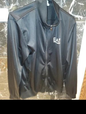 Svart ziptröja från Emporio Armani EA7 - Säljer en svart ziptröja från Emporio Armani EA7 med stilren design och logga på bröstet. Tröjan har lång ärm, ståkrage och hel dragkedja framtill. Perfekt för dig som gillar sportig och snygg streetstyle.