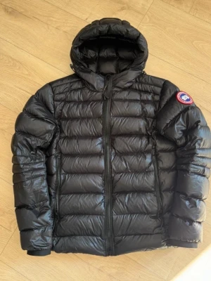 Svart dunjacka från Canada Goose - Säljer en svart, glansig dunjacka från Canada Goose med huva och klassisk logga på ärmen. Jackan har quiltad design, dragkedja framtill och två sidofickor. Perfekt för kalla dagar och har en riktigt snygg, modern look.