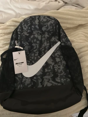 Svart och grå ryggsäck från Nike - Säljer en snygg ryggsäck från Nike med svart och grått kamouflagemönster och stor vit logga på framsidan. Väskan har meshfickor på sidorna och dragkedja upptill. Perfekt för dig som vill ha en cool och praktisk väska till skolan eller träningen.
