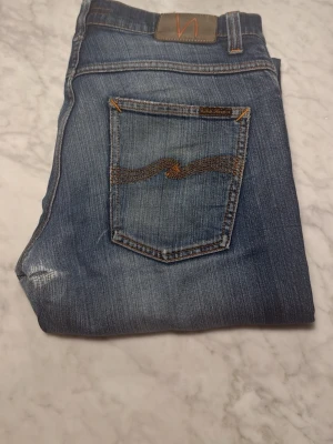Blå jeans från Nudie Jeans - Säljer ett par klassiska blå jeans från Nudie Jeans med snygga slitningar och kontrastsömmar. Modellen har raka ben och normal passform, med detaljerad brodering på bakfickan. Perfekt för dig som gillar en avslappnad och tidlös stil. (Väldigt efterfrågade och svåra att få tag på )