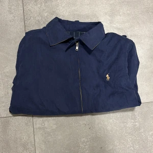 Marinblå vindjacka från Polo Ralph Lauren - Säljer en marinblå vindjacka från Polo Ralph Lauren med klassisk krage, dragkedja och broderad logga på bröstet. Jackan har rutigt innerfoder, elastiska muddar och två fickor framtill. Perfekt för dig som gillar stilren och tidlös design. Passar mer som S än M enligt mig.