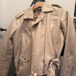 Beige skinnjacka från Zara med bälte - Snygg beige skinnjacka från Zara med coola dragkedjor, axelklaffar och bälte i midjan. Jackan har flera fickor med dragkedja och en klassisk bikerstil. Perfekt för dig som vill ha en stilren och trendig look.