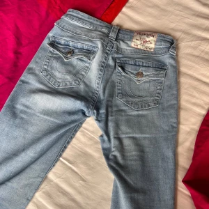 True Religion Becca Bootcut Jeans ljusblå - Ljusblå jeans från True Religion, modell Becca Mid Rise Bootcut. Snygga bakfickor med dekorativa sömmar och knappar. Klassisk bootcut-passform och tillverkade i mjukt denimtyg. Perfekta för dig som gillar en avslappnad men trendig look. Gick lite sönder mellan benen men syns inte mycket 