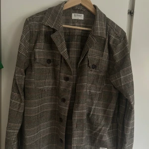 Rutig overshirt från Gabba - Snygg rutig overshirt från Gabba i grå och beige toner med bruna detaljer. Jackan har klassisk krage, knäppning framtill och två bröstfickor med knappar.