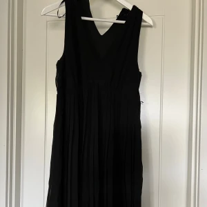 Svart klänning - Svart ärmlös klänning med v-ringning och plisserad kjol. Klänningen har dragkedja i sidan. 