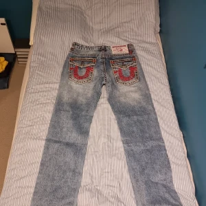 True Religion Joey jeans med brodyr - Säljer ett par True Religion Joey jeans i ljusblå. Bakfickorna har stor röd brodyr och logga, samt markerade knappar. Klassisk femficksmodell med breda sömmar och unik design. Har använt dom en hång då jag testade men insåg att de var för små i låren. Annars är de aldrig använda. Nypris 1600. Prisband finns kvar och originalpåse.