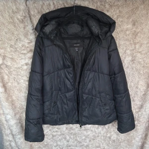 Svart pufferjacka med huva från Vero Moda - Svart pufferjacka från Vero Moda med stor, mysig huva och dragkedja framtill. 