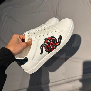 Gucci vita sneakers med orm brodyr - Säljer ett par vita sneakers från Gucci i skinn med klassiska gröna och röda ränder på sidan och en broderad röd-svart orm. Skorna har snörning, rund tå och platt sula. Perfekta för dig som vill sticka ut med en lyxig och trendig look.