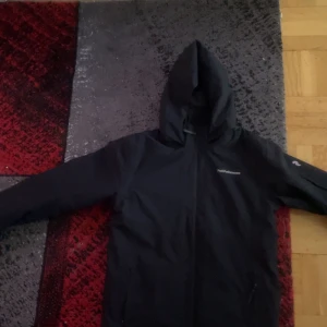 Peak performance jacka 170 - Svart vindjacka från Peak Performance i storlek 170 med huva och dragkedja framtill. Jackan har två sidofickor med dragkedja och en stilren design som passar perfekt för blåsiga dagar. Materialet är syntet och jackan har långärmade ärmar.