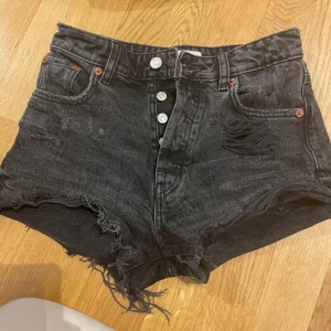 Svarta denimshorts från Zara - Snygga svarta denimshorts från Zara med hög midja, slitna detaljer och fransig kant. Klassisk femficksmodell med silverfärgade knappar framtill. Perfekta för en edgy och avslappnad stil i sommar.