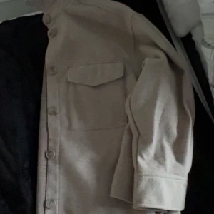 Beige kappa med fickor - Stilren beige kappa med två stora bröstfickor och knäppning framtill. Jackan har en rak passform och är tillverkad i ett mjukt material som känns skönt mot huden. Perfekt att slänga över en hoodie eller t-shirt för en avslappnad look.