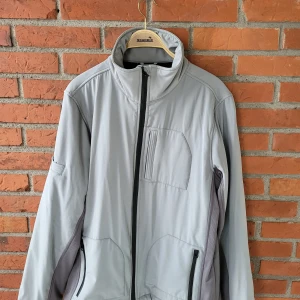 Mintgörn och grå vindjacka från Batistini L - Snygg ljusgrå vindjacka från Batistini i storlek L. Jackan har hög krage, svarta dragkedjor, flera fickor och coola detaljer på ärmen. Insidan är fodrad med mjuk svart fleece för extra komfort. Perfekt för dig som vill ha en stilren och funktionell jacka.