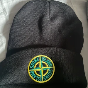 Svart Stone Island mössa med patch - Svart mössa från Stone Island med klassisk rund patch framtill i gult och grönt. Mössan är ribbstickad och har uppvikt kant för extra stilpoäng. Perfekt för dig som vill ha en clean look med street-vibe och ikonisk logga.