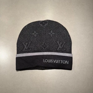 Svart LV mössa - Snygg svart mössa från Louis Vuitton med grå kant och ikoniskt LV-mönster över hela ytan. Märkets logga är broderad framtill och materialet känns mjukt och lyxigt. Perfekt accessoar för dig som vill ha en exklusiv look.