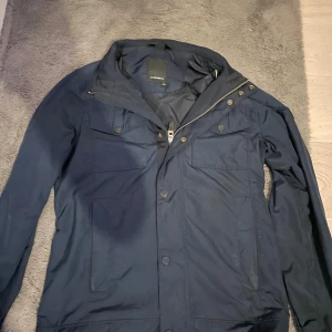 Mörkblå vindjacka från J.Lindeberg - Snygg mörkblå vindjacka från J.Lindeberg med huva, dragkedja och knappar framtill. Jackan har två bröstfickor med lock och två sidofickor. Perfekt för dig som vill ha en stilren och funktionell jacka till vardags.