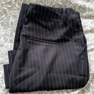 Pw Wilma 42 - Snygga svarta kostymbyxor med tunna vita ränder i klassiskt randigt mönster. Byxorna har raka ben, bälteshällor och en bakficka. Modell pw Wilma från bikbok, helt nya och tror inte denna färg säljs mera