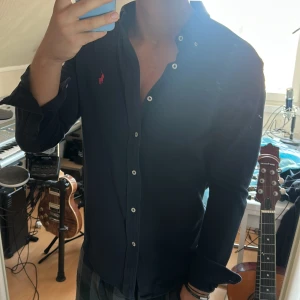 Mörkblå skjorta från Ralph Lauren - Snygg mörkblå skjorta från Ralph Lauren med klassisk röd broderad logga på bröstet. Skjortan har lång ärm, vita knappar och en normal passform. Tillverkad i mjuk bomull som känns skön mot huden. Perfekt för dig som gillar stilrena och tidlösa plagg.