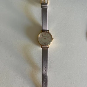 Olivia Burton klocka med grått läderband - Stilren klocka från Olivia Burton med rund urtavla i vitt och guldfärgad ram. Klockan har romerska siffror och ett grått läderband som ger en klassisk och elegant look. Perfekt accessoar för dig som gillar enkel men snygg design.