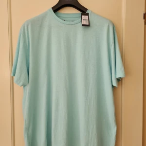 Ljusblå basic t-shirt från Dressmann strl 3XL - Enkel och stilren ljusblå t-shirt från Dressmann i storlek 3XL. T-shirten har rund hals, korta ärmar och är tillverkad i en blandning av bomull och polyester.
