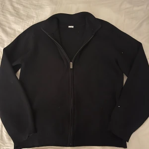 Svart stickad kofta med dragkedja - Svart stickad kofta med hel dragkedja framtill och ribbad krage. Långärmad modell som är enkel att matcha med olika outfits. Perfekt för kyliga dagar när du vill ha något stilrent och bekvämt. Stickat material 