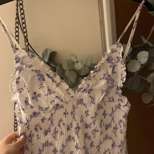 Vit Zara blommig kortklänning med volang - Supersöt populär Zara vit kortklänning  med blått och lila blommönster, smala axelband och volangdetaljer vid bysten. Klänningen har smockad överdel och lätt utställd kjol, perfekt för varma dagar. Materialet känns lätt och luftigt. Materialet drar sig efter midjan som gör att det ser ännu bättre ut 
