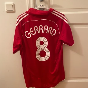 Liverpool Gerrard 8 Adidas tröja M - Röd Liverpool fotbollströja från Adidas med Gerrard och nummer 8 på ryggen. Klassisk design med vita detaljer, Carlsberg-logga på bröstet och Champions League-märke på ärmen. Tillverkad i lätt och ventilerande material, perfekt för match eller träning.  (Helt ny)